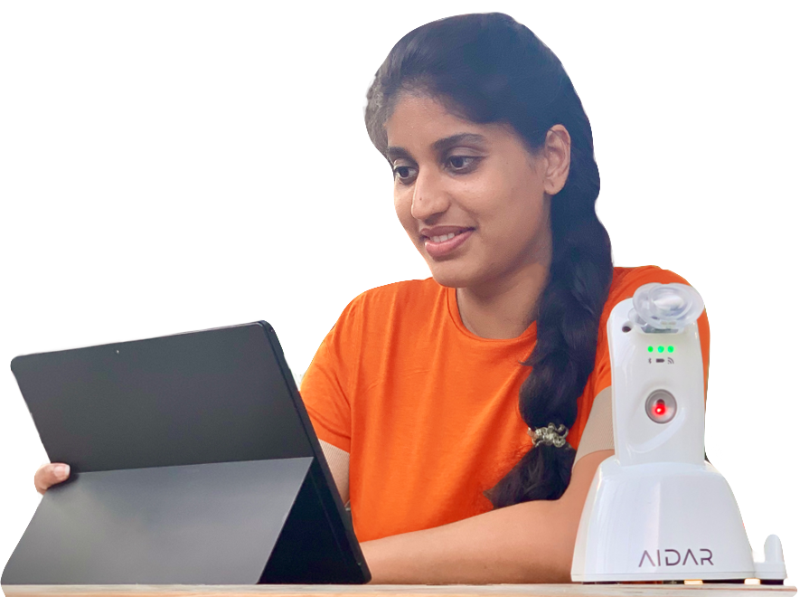 A young woman in an orange shirt on a telehealth video visit on her tablet at home, with a MouthLab device resting in its AIDAR charging dock beside her.